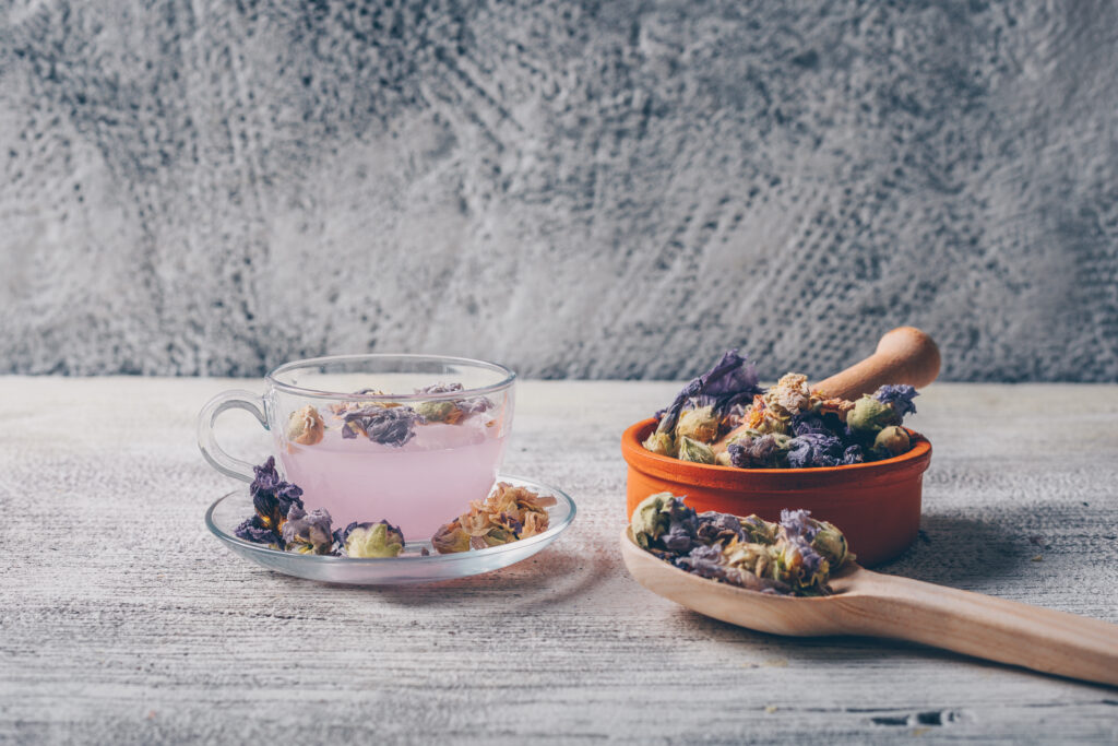 purple colored water in a cup with dried flower tea in a bowl and spoon side view on a white wooden and gray background free space for your text